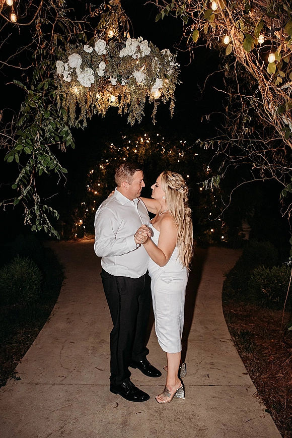 First dance under string lights as bride in strapless white dress and groom in white shirt sway beneath a floral chandelier on a garden path at night