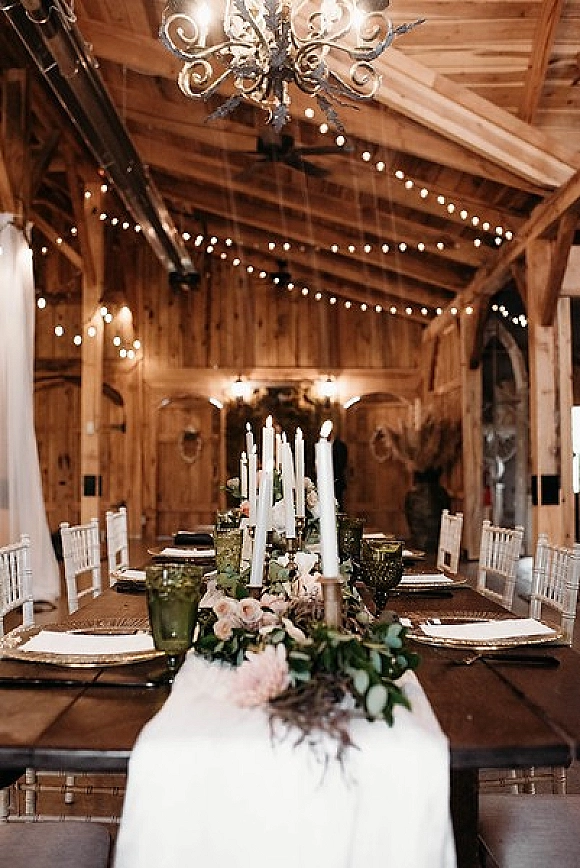 Reception tablescape with rustic wedding table decor, taper candles and floral greenery runner on a long farm table beneath chandeliers in a barn