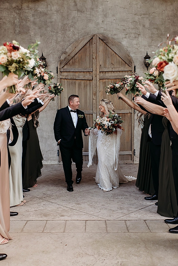 Wedding recessional as newlyweds walk out holding hands, bride in lace sleeves with bouquet, bridesmaids cheer by wooden doors in courtyard