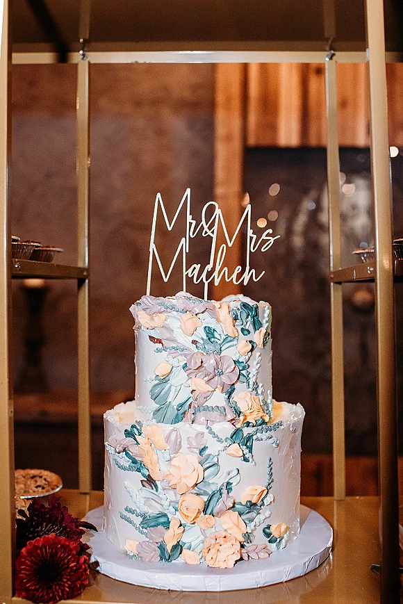 Wedding cake, two tier wedding cake with hand-painted floral icing and cake topper on a gold stand, burgundy flowers and warm bokeh lights