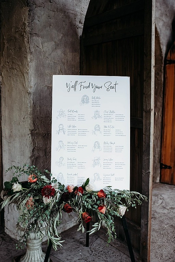 Wedding seating chart on an easel with a seating chart sign and floral greenery urn beside a stone wall and arched wooden doorway