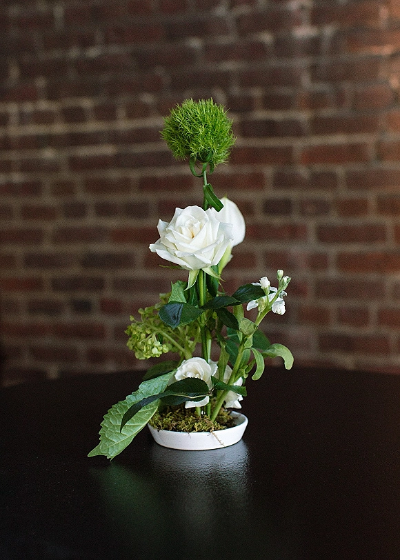 Wedding centerpiece with white roses and a single green stem nestled in moss in a white ceramic dish on a dark table before a brick wall
