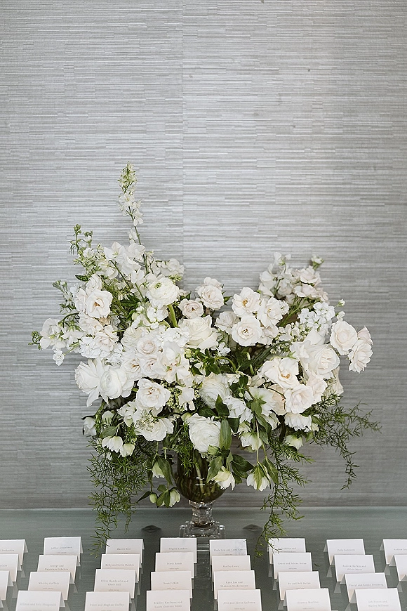 Wedding escort cards on an escort card table with a white floral arrangement in a glass vase and greenery against a gray textured wall