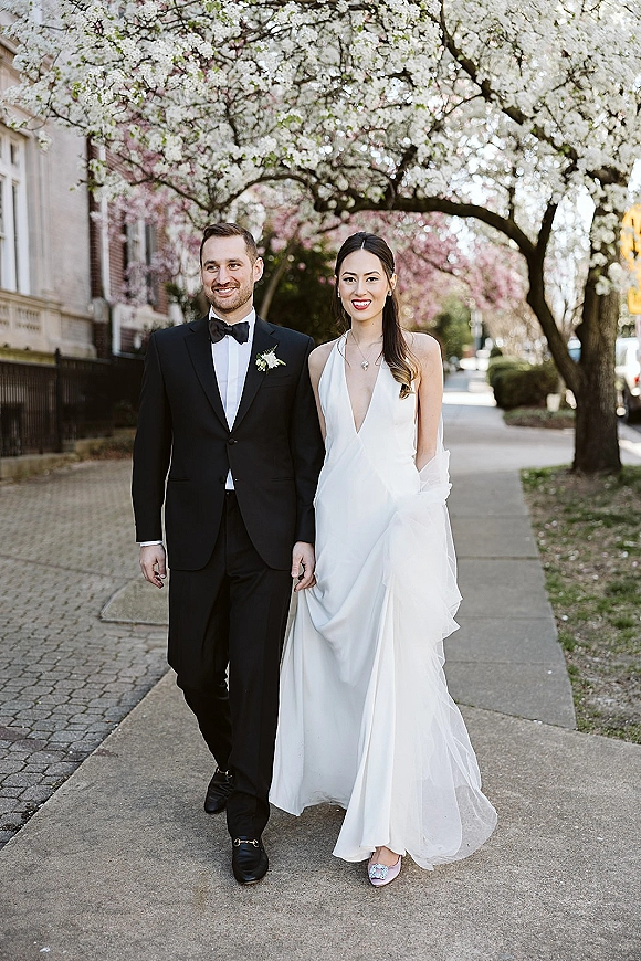 Couple portrait of bride and groom walking hand in hand, her deep V wedding dress and his tuxedo beneath flowering trees on a city sidewalk