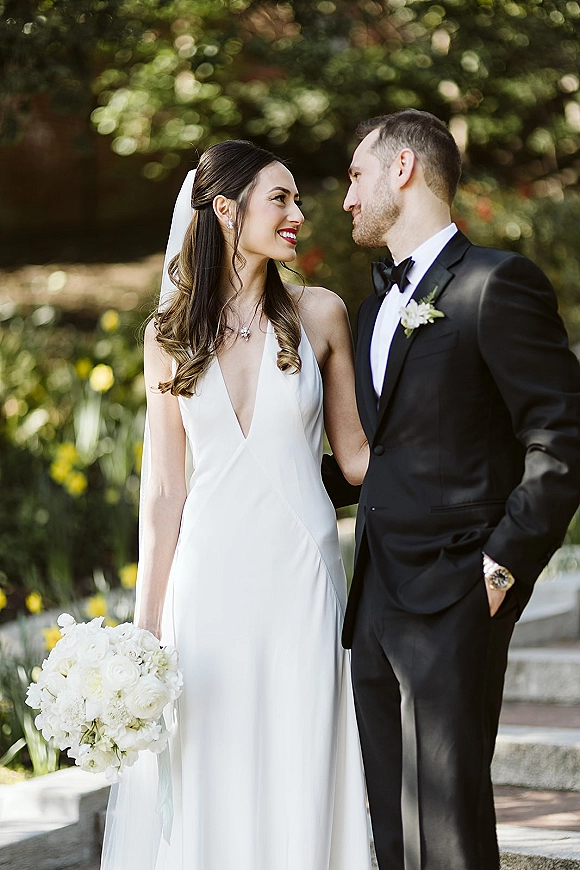 Couple portrait of bride and groom looking at each other, bride holding a white rose bouquet, in sunlit garden by stone steps