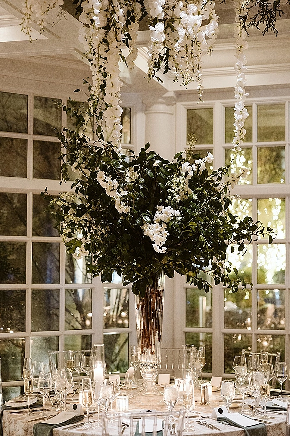 Reception tablescape with tall floral centerpiece of white blooms and greenery, taper and votive candles, set before large grid windows