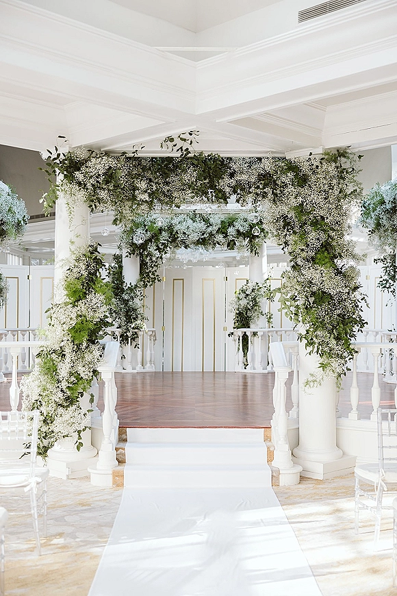 Ceremony backdrop with a greenery wedding arch and baby’s breath, hanging glass orbs, and a white aisle runner in a bright indoor venue
