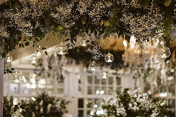 Hanging floral installation with baby’s breath and greenery, dotted with glass votives and candles beneath tall reception windows