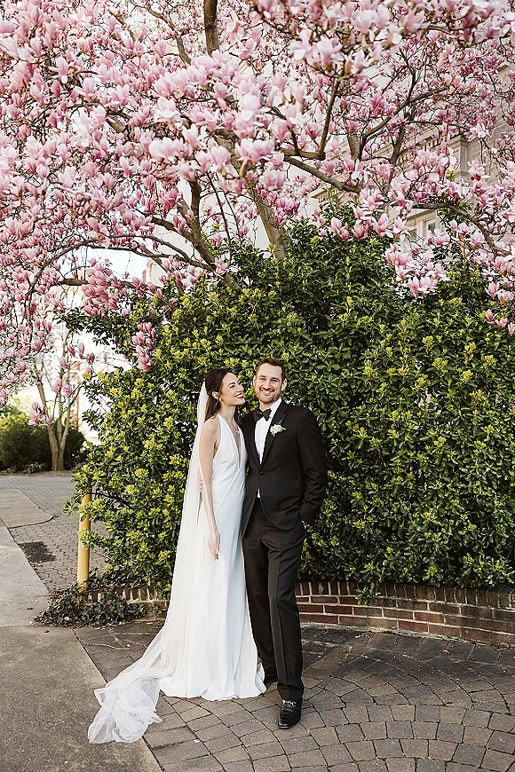 Couple portrait of bride in satin wedding dress and veil leaning on groom in black tux, smiling beneath a pink flowering tree by a brick wall