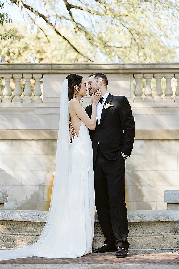 Wedding couple portrait of bride touching groom’s face in a kiss pose, long veil flowing by a sunlit stone balustrade in a garden