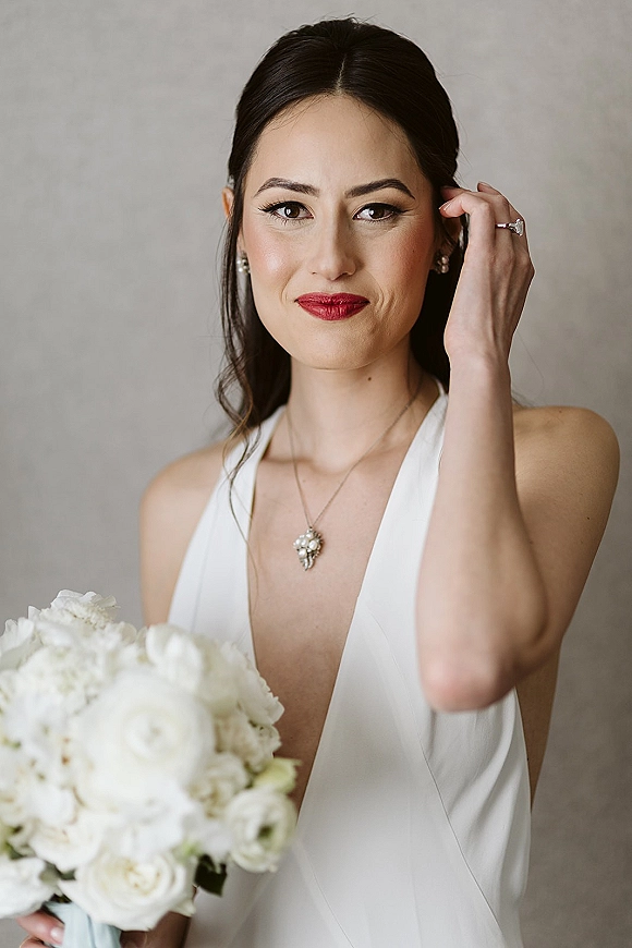 Bridal portrait of a bride holding bouquet of white roses, wearing a halter wedding dress and red lipstick against a neutral wall
