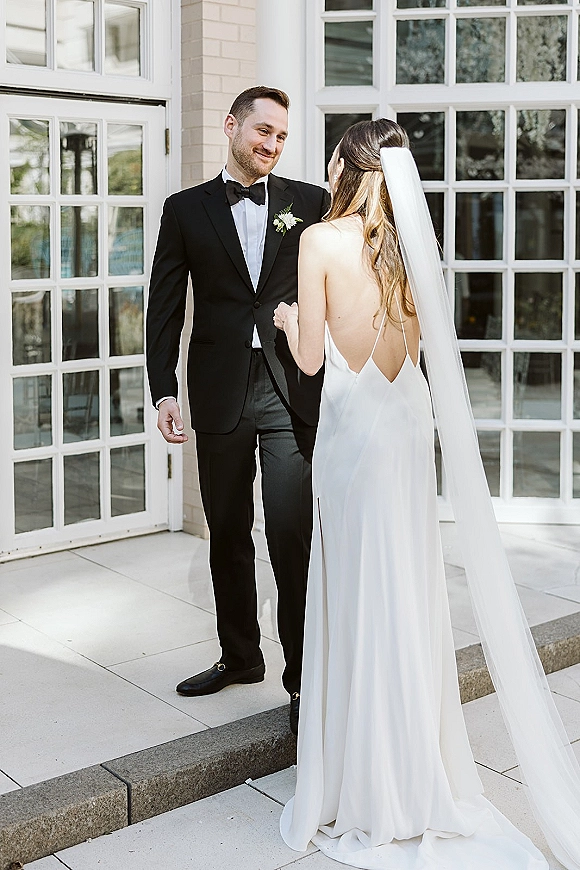 First look moment as bride in a wedding dress and long veil meets groom in black tuxedo at white French doors on stone steps