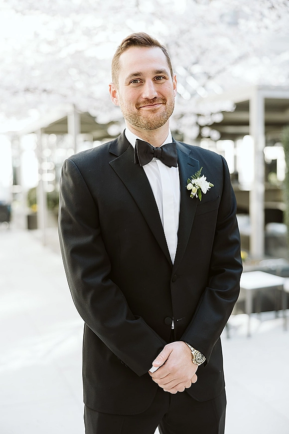 Groom portrait in a black tuxedo with bow tie and white boutonniere, hands clasped on an outdoor walkway by blooming trees and columns