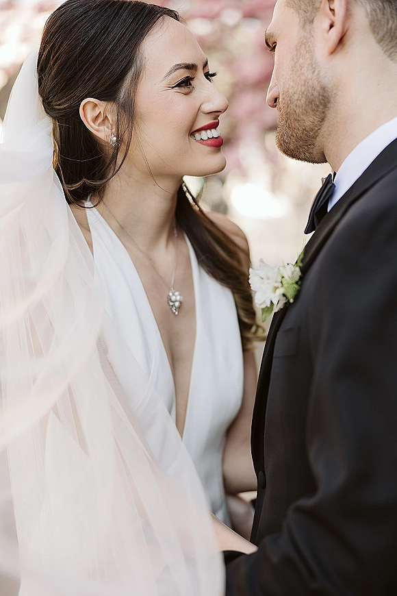 Wedding couple portrait with bride smiling at groom, foreheads touching under her veil, blurred greenery and pink blossoms behind them
