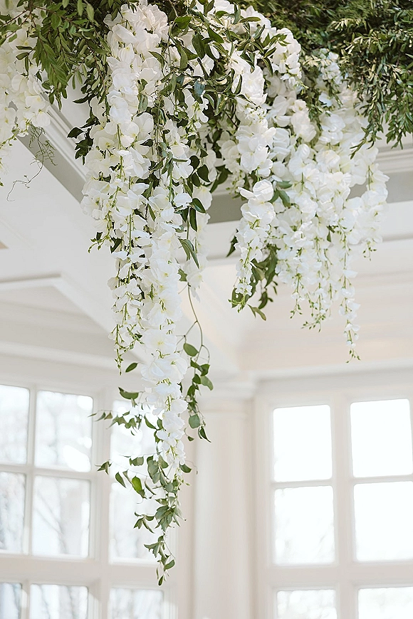 Hanging floral installation with ceiling floral decor of white blooms, greenery garland, and trailing vines beneath large windows