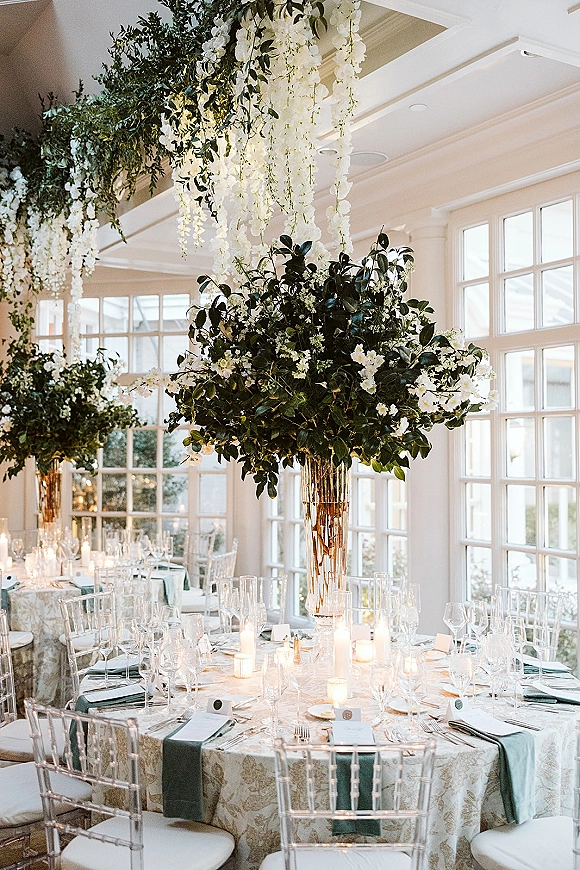 Reception tablescape with round wedding table decor, tall floral centerpieces, taper candles, and greenery under hanging white florals in a sunroom