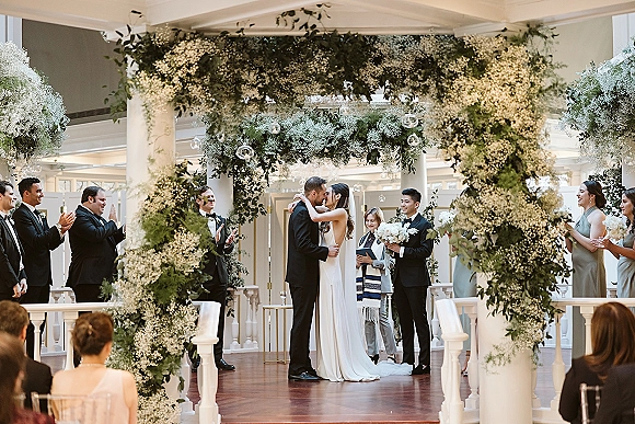 Ceremony kiss as bride in veil and groom in tuxedo embrace beneath baby’s breath floral arch with hanging glass orbs indoors