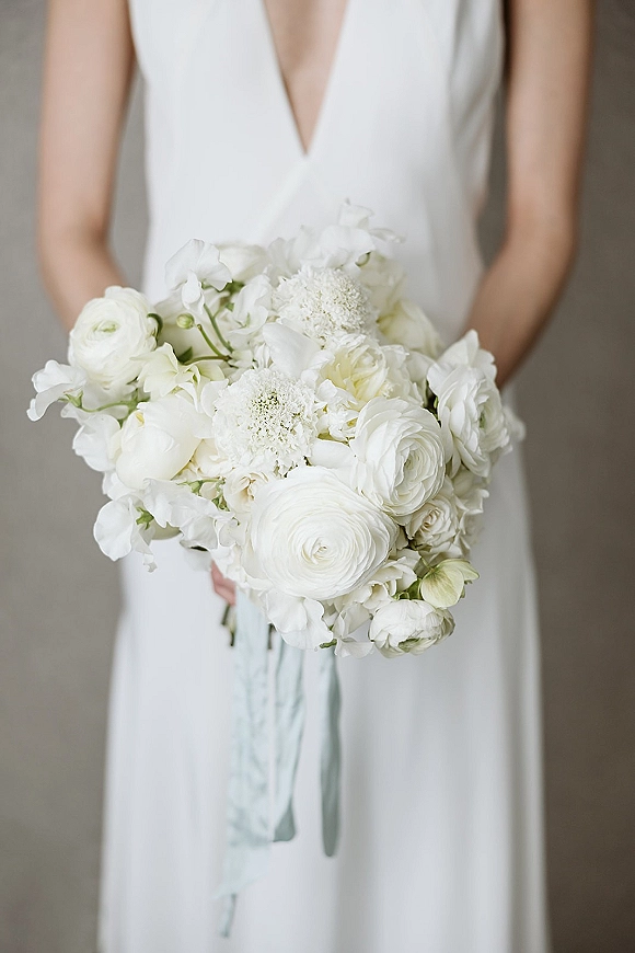 Bridal bouquet of white flowers with ranunculus and sweet peas, tied with a pale blue ribbon against a neutral wall backdrop