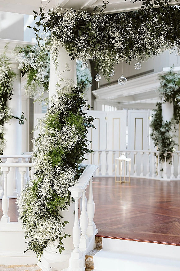 Wedding ceremony decor with ceremony arch greenery, baby's breath garlands and hanging glass globes on white columns by a staircase