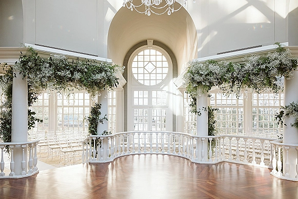 Indoor ceremony setup with a wedding ceremony arch draped in greenery garland beneath a chandelier, facing a grand window wall and white chairs