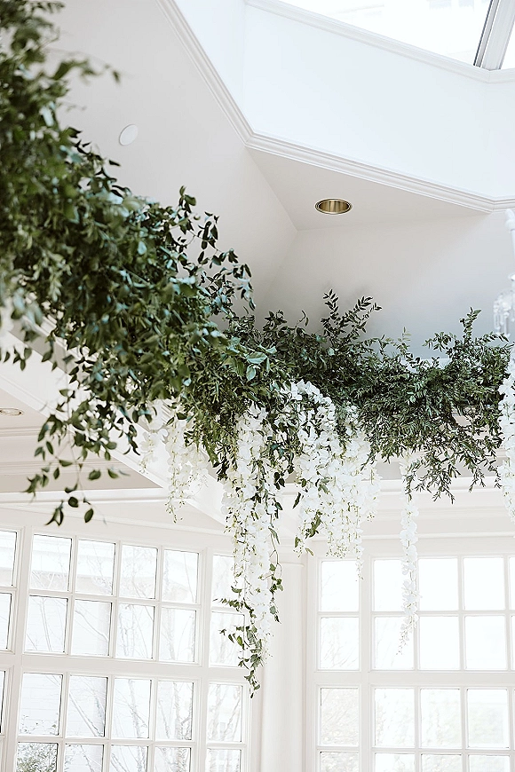 Hanging floral installation with wedding ceiling greenery and white wisteria strands under a skylight ceiling and large windows backdrop