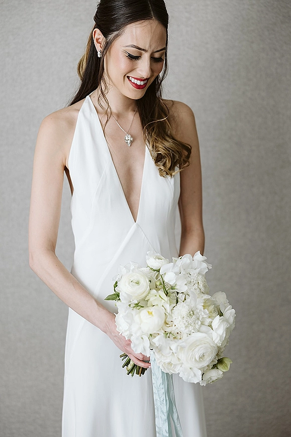 Bridal portrait of a bride holding bouquet of all-white flowers with ribbon wrap, red lipstick and jewelry against a neutral wall
