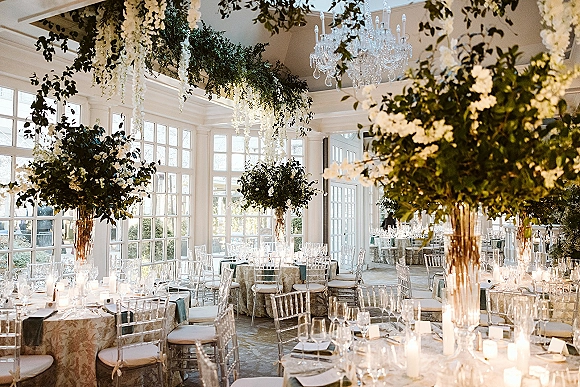 Reception tablescape with hanging floral installation, tall white centerpieces in glass vases, candles and crystal chandelier beside window wall
