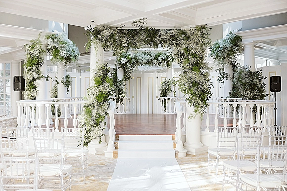 Ceremony setup with an indoor wedding ceremony floral arch, clear chiavari chairs and white aisle runner beneath hanging glass orbs in a bright venue