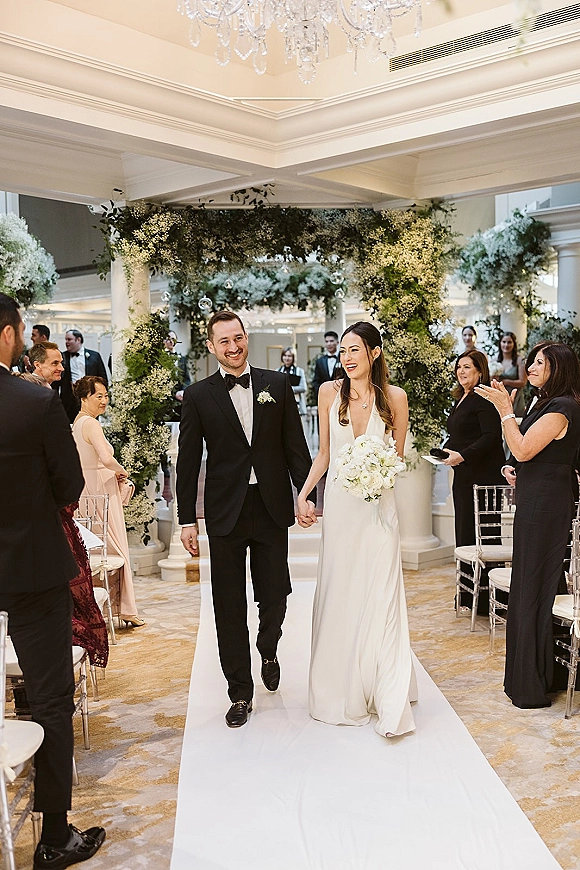 Wedding recessional with bride and groom walking down aisle holding hands, bride with white bouquet under chandeliered ballroom arch