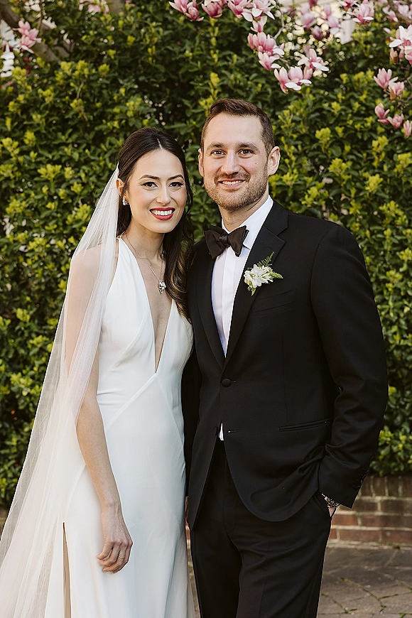 Couple portrait of bride in a long veil and white gown with groom in a black tuxedo, posed by a hedge and pink magnolias