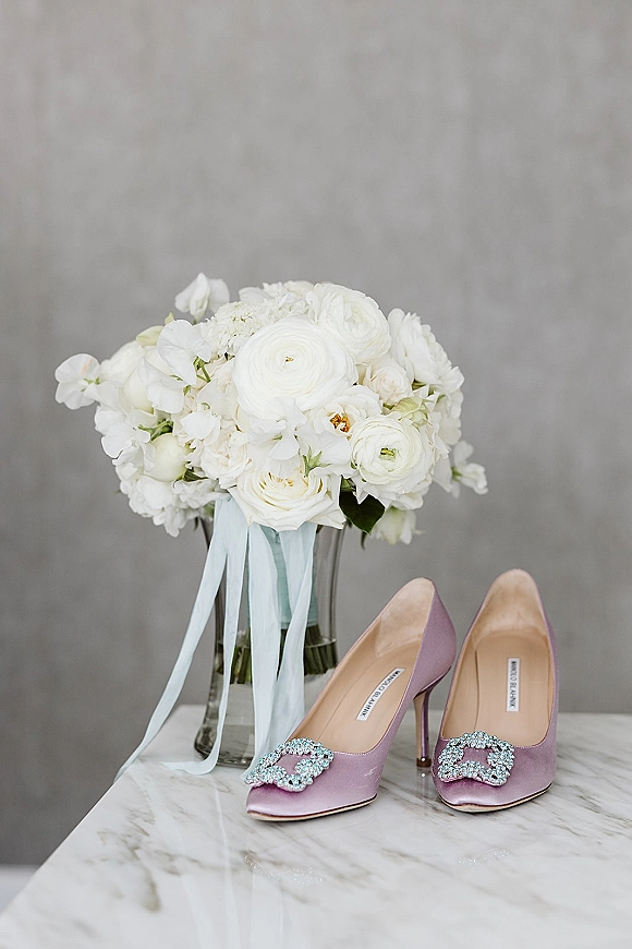 Bridal shoes in lavender wedding heels with a crystal buckle beside a white ranunculus bouquet and ribbon on a marble tabletop