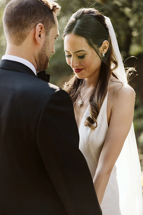 Wedding couple portrait with bride looking down in a long veil and white dress beside groom in tuxedo, framed by lush trees outdoors