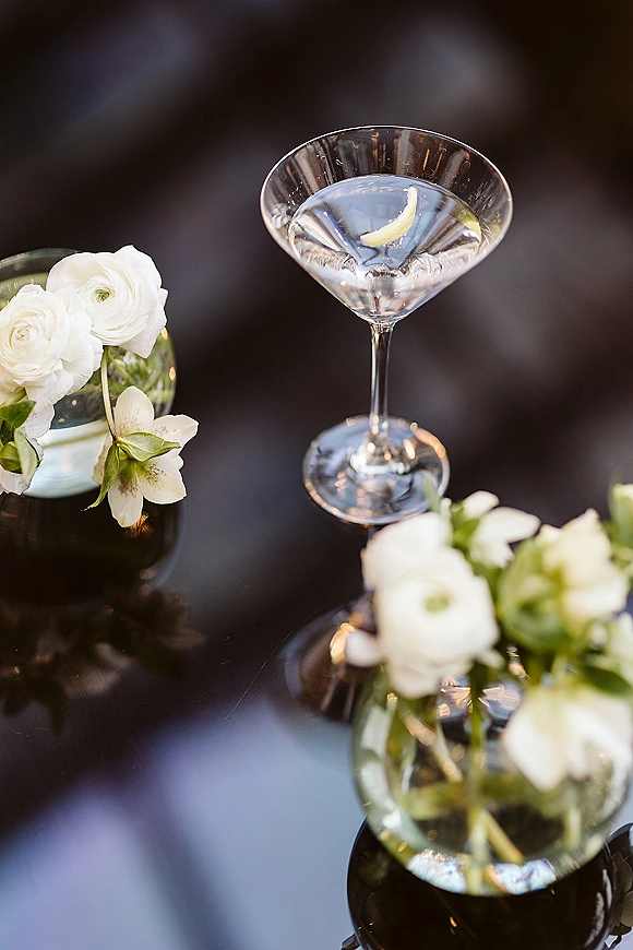 Signature wedding cocktail in a martini glass with lemon twist garnish beside white flowers in bud vases on a glossy black tabletop