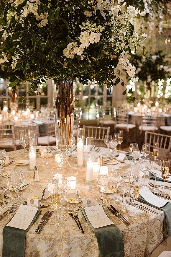 Reception tablescape with wedding table centerpiece of white flowers and greenery in a glass trumpet vase, taper candles under string lights