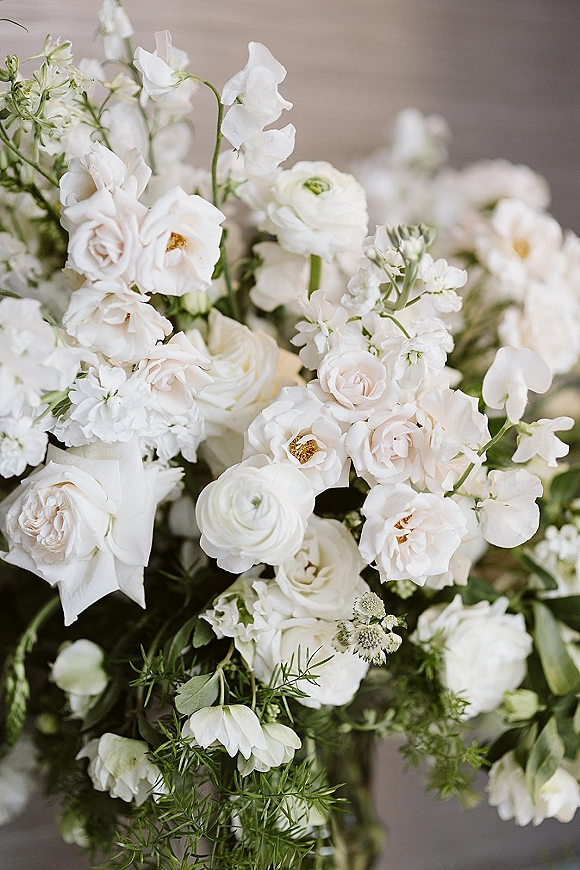 Bridal bouquet of white roses and ranunculus with sweet peas, white blossoms, and greenery, tied with ribbon in soft indoor light