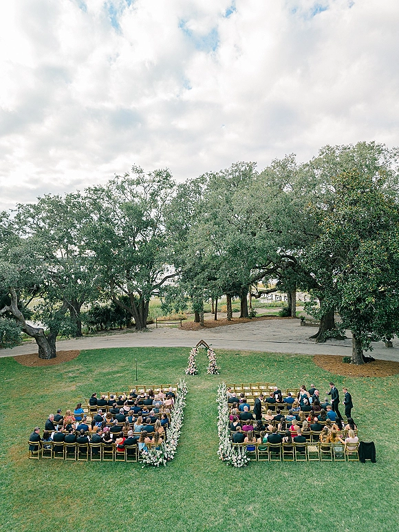 Ceremony setup for an outdoor wedding ceremony with a floral arch and aisle flowers, wooden chairs on a grassy lawn beneath large trees