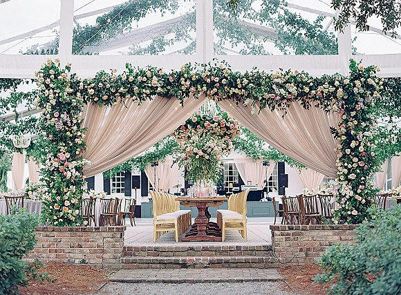 Wedding reception decor with a floral arch backdrop framing a long wooden table with gold chairs, candles, and rose greenery garlands under a white pavilion