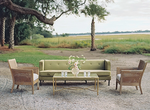 Wedding lounge seating with a green velvet sofa and cane-back chairs around a glass coffee table on a coastal lawn by water under clouds