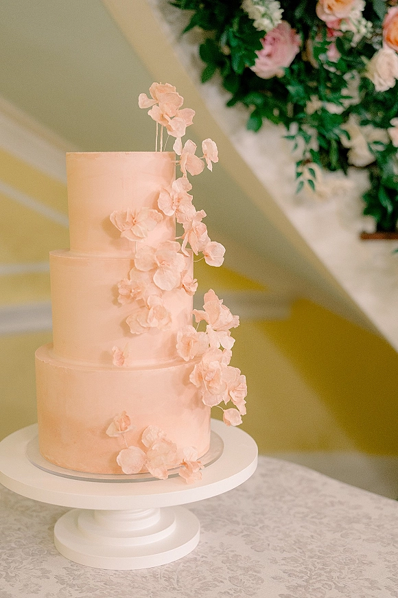 Wedding cake with blush pink frosting and cascading sugar flowers on a white stand atop a lace tablecloth near a staircase