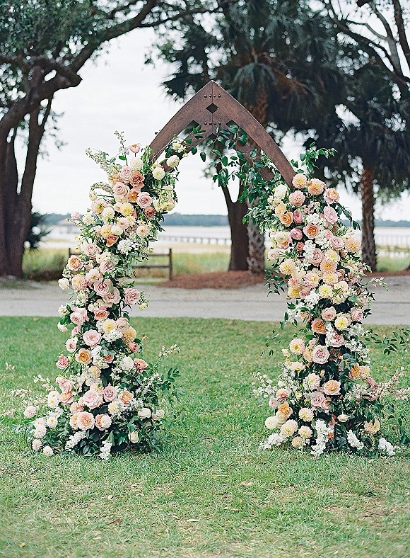 Wedding ceremony arch with a lush floral arch of roses and dahlias on a metal frame, set on a lawn by palm trees and waterfront