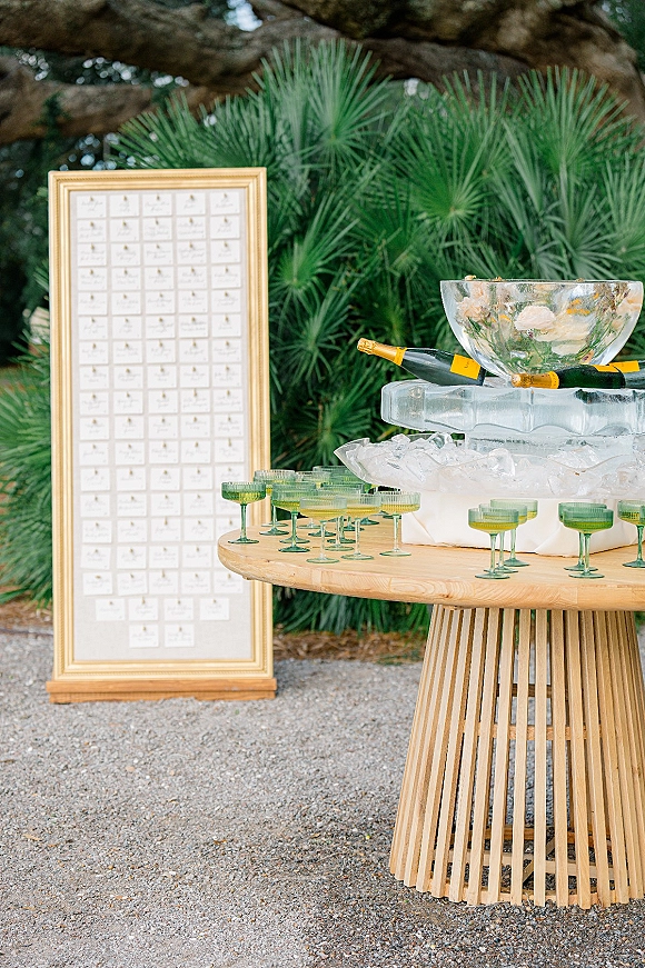 Wedding escort display with escort card wall in a gold frame on a wooden stand, beside champagne bottles on ice and coupe glasses under palms outdoors