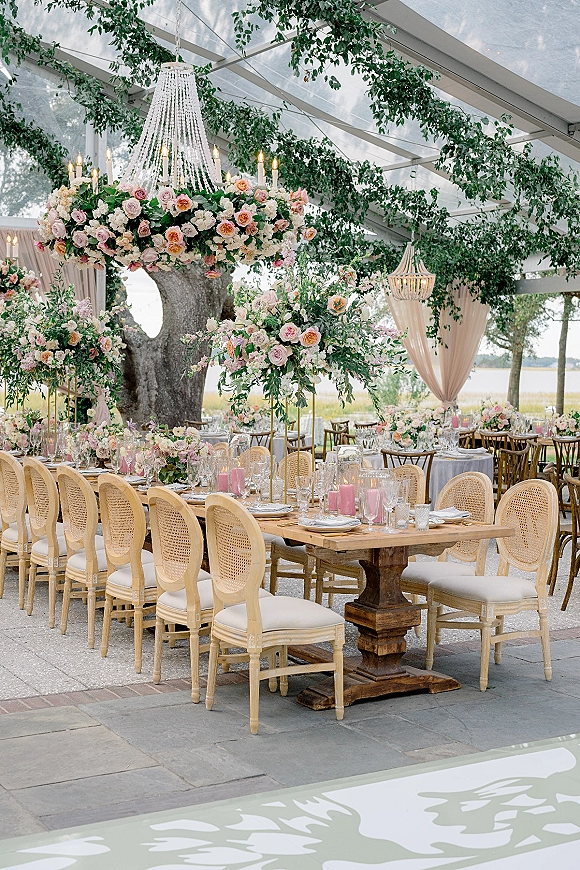 Reception tablescape with floral centerpieces and greenery garlands under a clear top tent, candlelit farm tables by a waterfront view