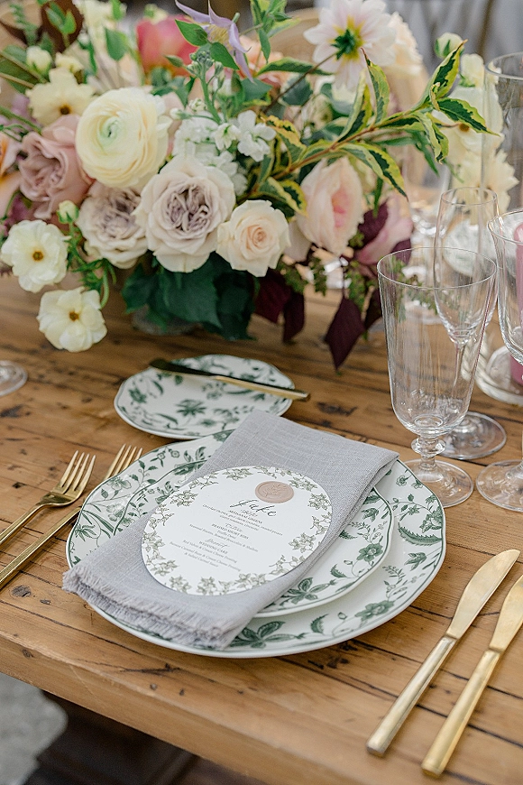 Reception tablescape with wedding place setting, blush rose centerpiece and greenery, patterned plates, wax-sealed menu, and gold flatware on a wooden table