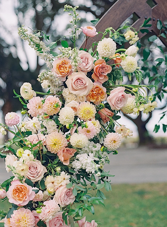 Wedding floral arrangement with ceremony arch flowers in peach and blush roses, dahlias, and greenery on a wooden structure outdoors