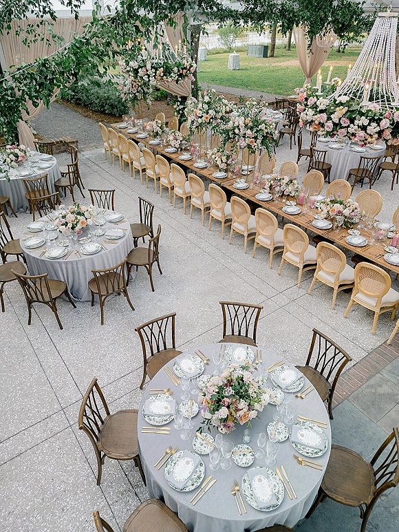 Wedding reception decor with long banquet and round tables in gray linens, floral garlands and candles under chandeliers on a lakeside patio