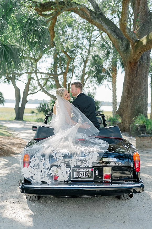 Couple portrait in a vintage convertible, bride and groom in car looking back, her long veil flowing on a waterfront dirt road