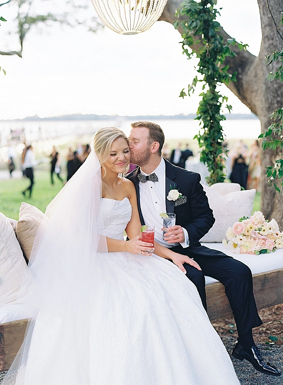Wedding couple portrait of groom kissing bride’s cheek as they sit on a lounge sofa holding cocktails, with chandelier and waterfront lawn backdrop
