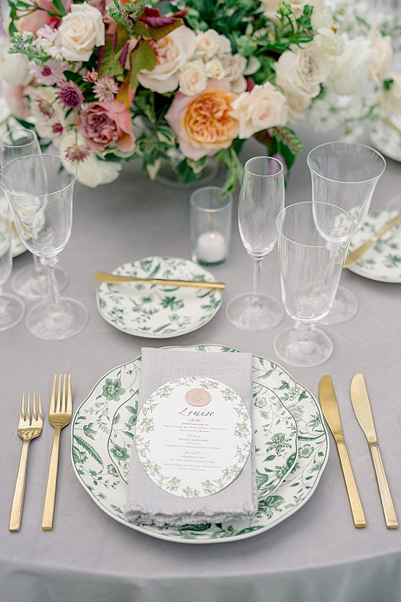 Reception tablescape wedding place setting with botanical plates, gold flatware, grey linen napkins, menu card, and rose centerpiece on round tablecloth
