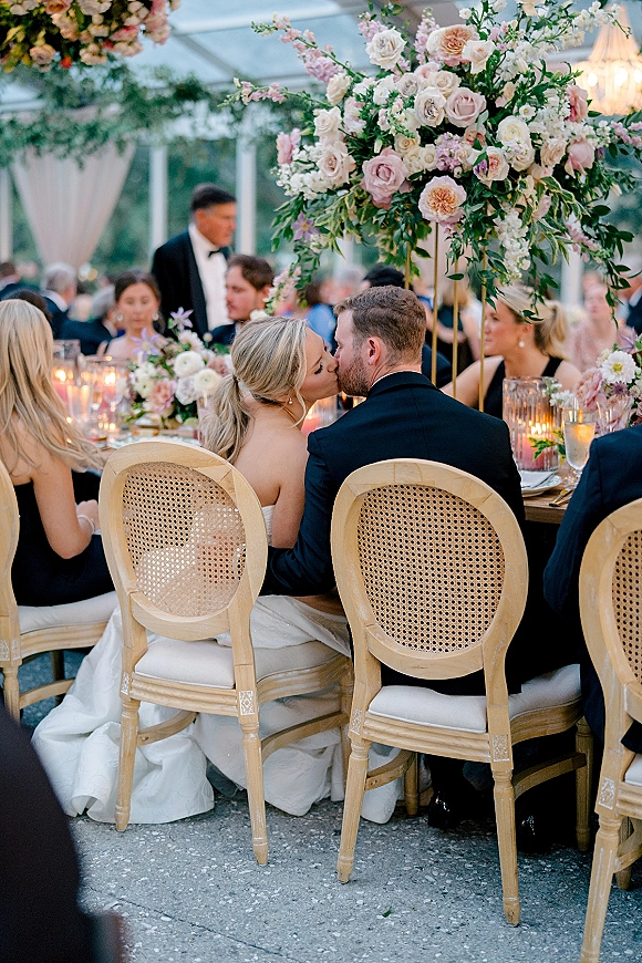 Wedding kiss at the reception as bride in a strapless gown and groom in a black tuxedo lean in at a candlelit sweetheart table under string lights