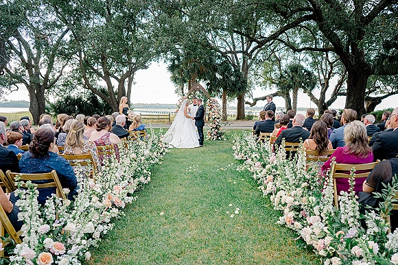 Wedding ceremony with bride and groom at altar beneath a floral arch, guests seated on lawn by waterfront pier under oak trees